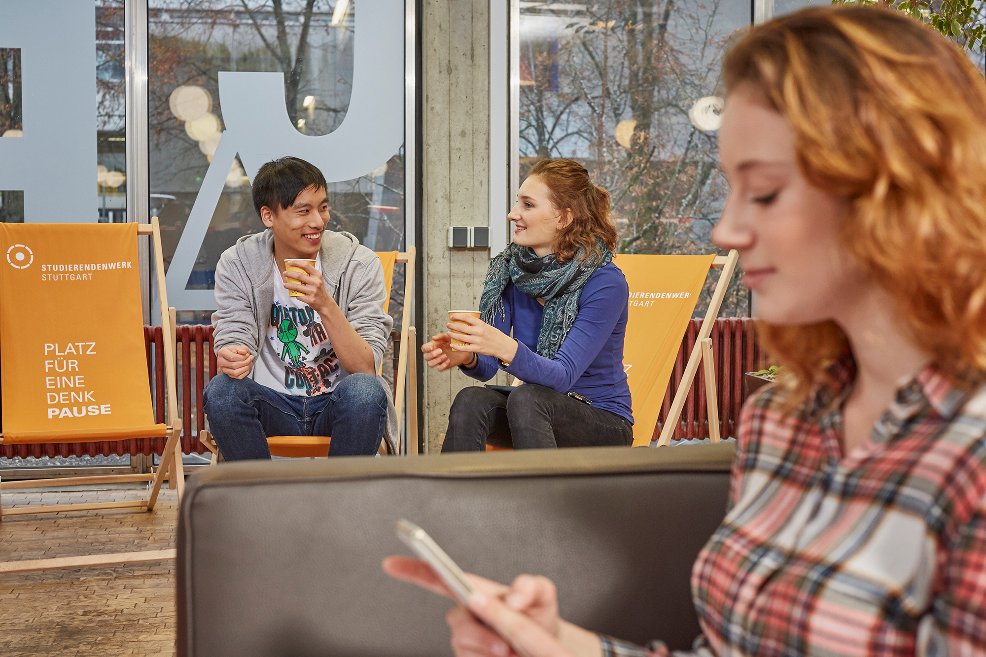 Close-up of a student in the coffee lounge in the cafeteria in Vaihingen. In the background, two other students are talking.