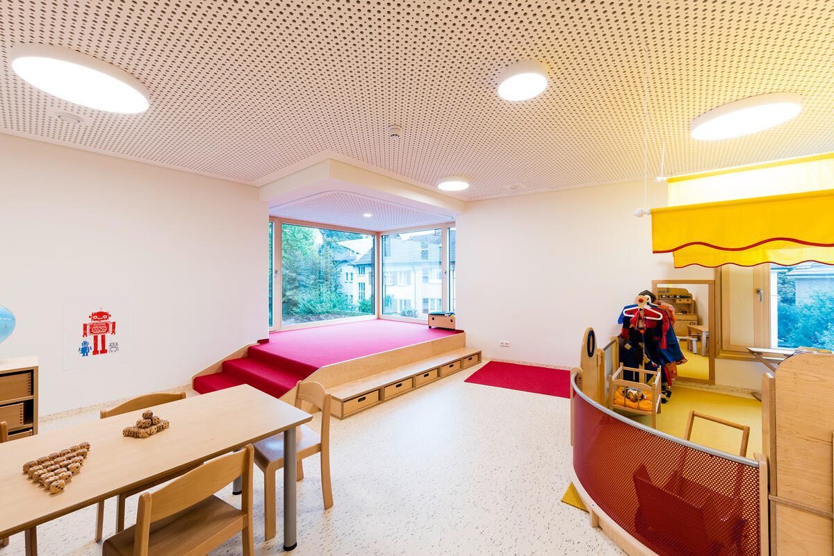 Group room with table and chairs and play corner with red carpet in Kita Abenteuerbande