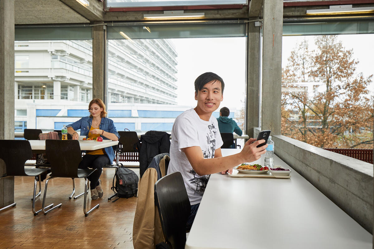 Student sitzt im Speisesaal beim Essen und spielt am Handy