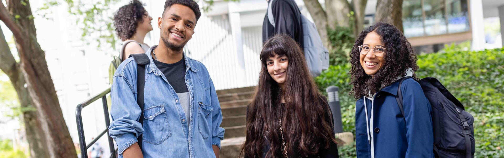 A male and two female students stand on the university campus and smile at the viewer.