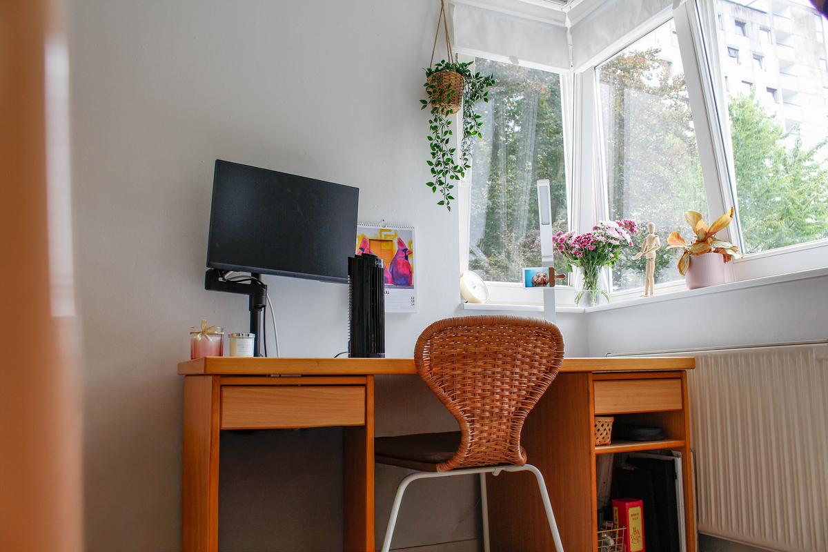 Schreibtisch mit Bildschirm und einem Stuhl. Rechts ist ein Fenster zu sehen