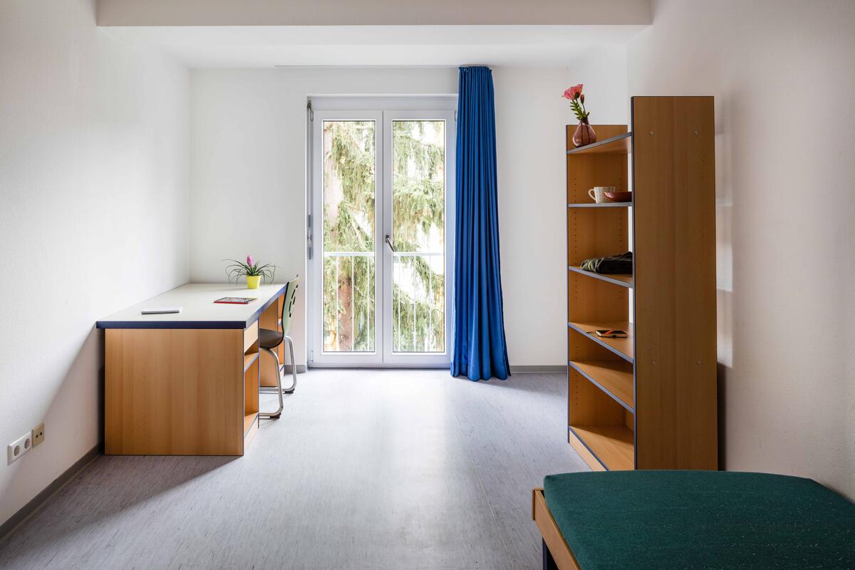 Zimmer mit Schreibtisch, Bett, Regal und großem Fenster im Studentendorf Göppingen