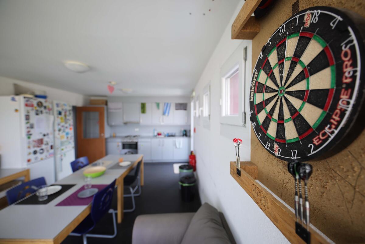 Küche mit langem Tisch, Stühlen und einer Dartscheibe an der Wand Zimmer mit Bett, Schreibtisch und Regal im Wohnheim in der Fabrikstraße