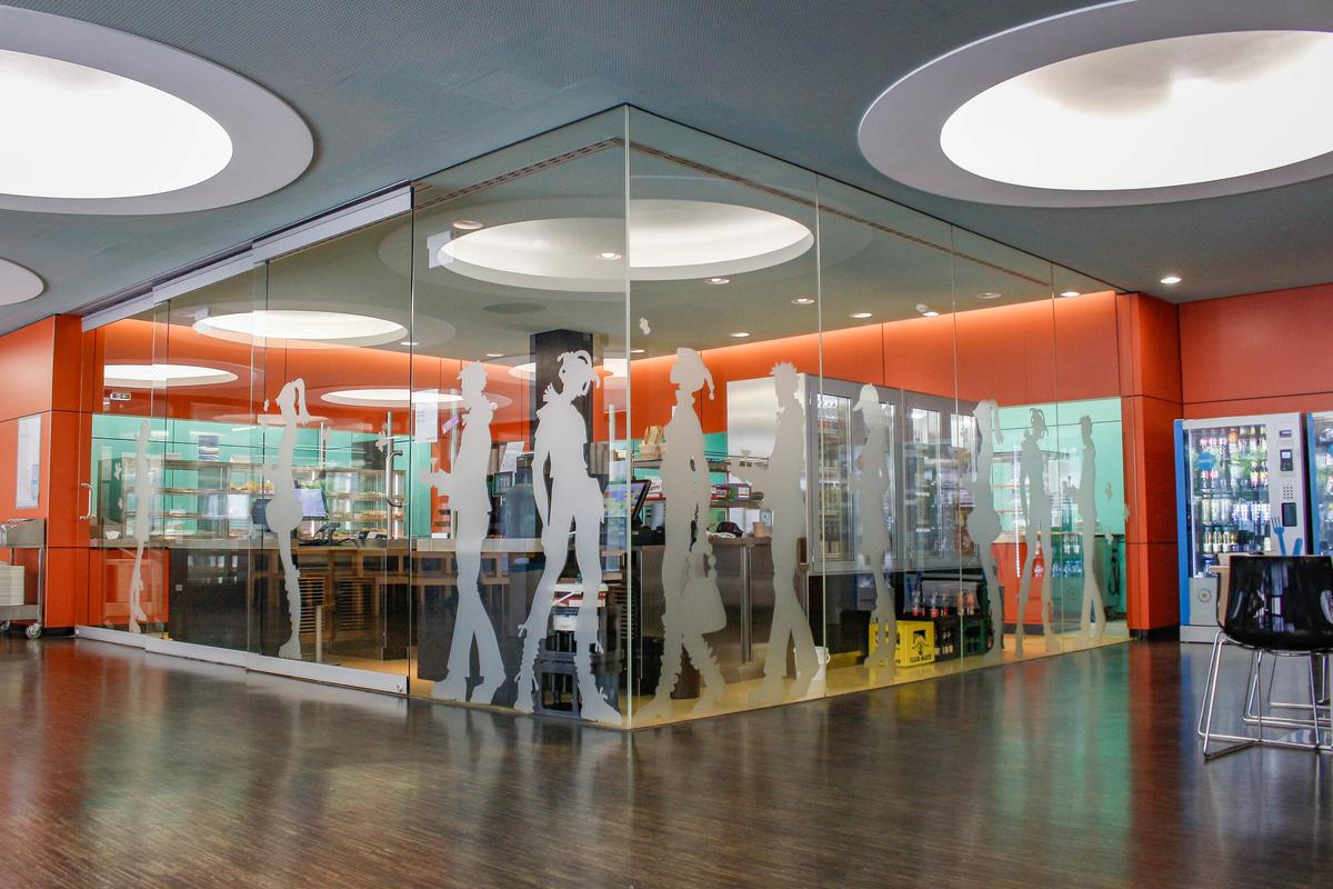 Cafeteria Lichtblick. Die Snackausgabe ist umgeben von Deckenhohen Scheiben auf denen Silhouetten von Personen foliert sind.